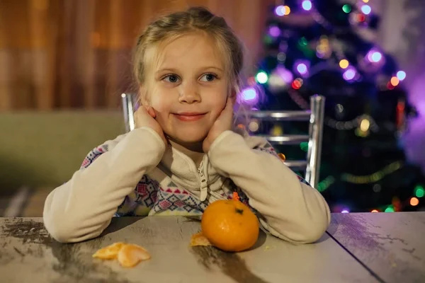 Une fille aux cheveux bruns nettoie une mandarine assise à une table — Photo