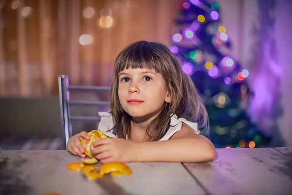 Une fille aux cheveux bruns nettoie une mandarine assise à une table — Photo