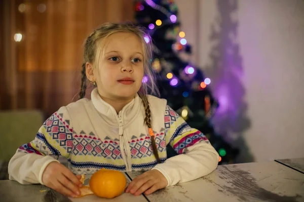 Une fille aux cheveux bruns nettoie une mandarine assise à une table — Photo