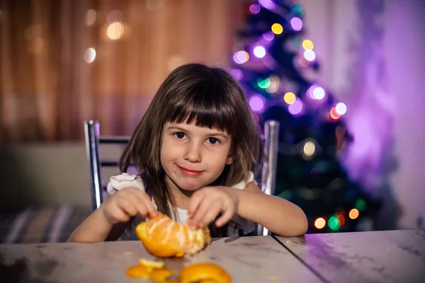 Une fille aux cheveux bruns nettoie une mandarine assise à une table — Photo
