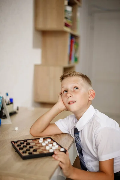 一个穿着白衬衫的男孩坐在桌子前玩跳棋游戏. — 图库照片