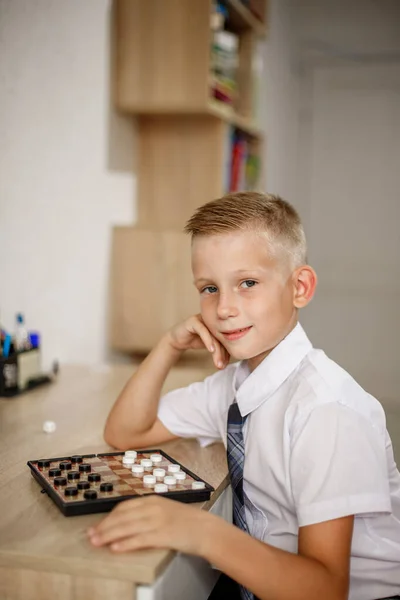Ένα αγόρι με λευκό πουκάμισο παίζει ντάμα ενώ κάθεται σε ένα γραφείο.. — Φωτογραφία Αρχείου