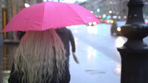 Girl walking under an pink umbrella in the city center in the rain. Blonde girl under an umbrella walking down the street