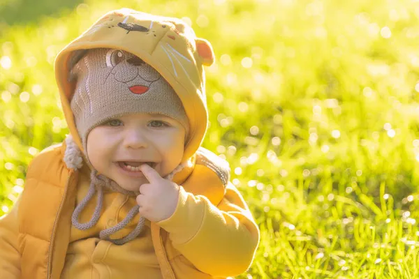 El niño de un año se sienta en la hierba verde y llora. Niño molesto con sombrero divertido y chaqueta amarilla. Retrato de un niño ofendido en el parque de otoño. — Foto de Stock