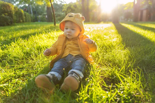 Retrato de un lindo niño sentado en la hierba verde al aire libre en el parque de otoño al atardecer. Un niño camina en el parque al aire libre. Feliz verano y concepto de estilo de vida. Infancia. — Foto de Stock