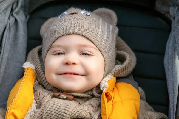 Pequeño niño alegre en el cochecito. Primer plano retrato de niño lindo usando sombrero de punto divertido y suéter caminando en el parque — Foto de Stock