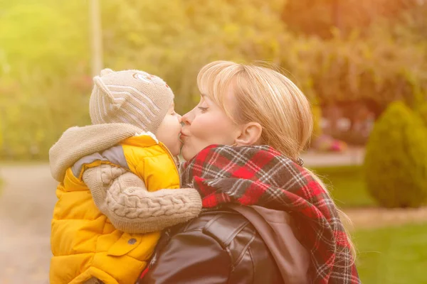 Mamá divirtiéndose con su hijo pequeño en el parque de otoño al atardecer. Mamá besa a su pequeño bebé, de cerca. Amor y cuidado. Concepto de familia feliz. — Foto de Stock