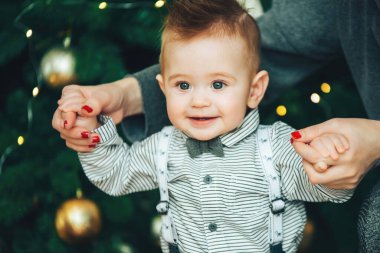 Çocuğun ilk Noel 'i. Noel ağacına karşı duran sevimli küçük çocuk. Yeni yıl konsepti. Sevimli çocuk Noel 'in tadını çıkarıyor. Anne küçük bir oğlanın elini tutuyor.