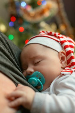 Annesinin kollarında uyuyan bir bebeğin portresi süslü bir Noel ağacının arka planına karşı. Olay, Noel ve Yeni Yıl. Çizgili cüce şapkalı şirin noel bebeği.