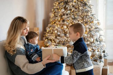 Küçük çocuk Noel hediyesiyle annesini kucağında bebekle karşılıyor. Mutlu aile - kardeşler ve anne Noel 'i ya da yeni yılı kutlar. Zarif ağacın yanındaki beyaz koltukta oturan kadın..