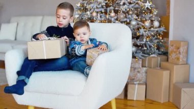 Ellerinde hediyelerle rahat koltukta oturan iki neşeli çocuk. Noel Baba 'nın hediyesi. Neşeli ruh hali. Noel sabahı aydınlık oturma odasında. Yavaş çekim