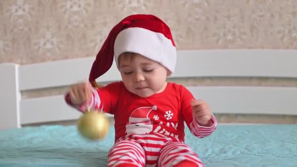 サンタの帽子の面白い小さな白人の赤ちゃんは 1本の指の黄金のクリスマスボールと笑顔でねじれています サンタ ベイビー 冬の休日やお祝いのコンセプトで家を準備し 飾る家族 — ストック動画