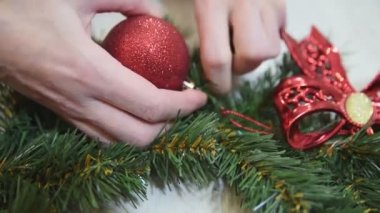 Kapalı kadın elleri bir Noel çelengini kırmızı parlak topla süslüyor. El işi yapmak. Mevsimlik tatil atölyesi çevrimiçi, evde şenlik dekorasyonu yapıyor. Seçili odak.