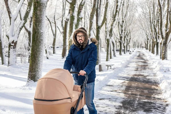 Kaukázusi férfi, aki buggyval sétál. Szakállas apa babakocsit tologatott a téli parkban a hóvihar után. Csecsemővel tölteni az időt egy gyönyörű, hideg napon. Stock Fotó