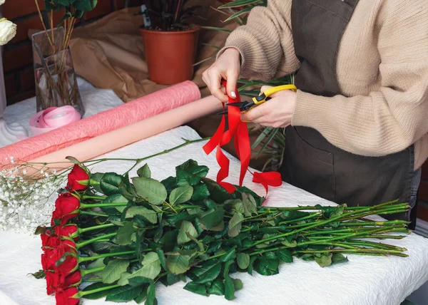Florist at work. Close-up female hands cut a colored ribbon. Woman making a bouquet of red roses for valentines day. Fresh roses for bouquet delivery. — Stock Photo, Image