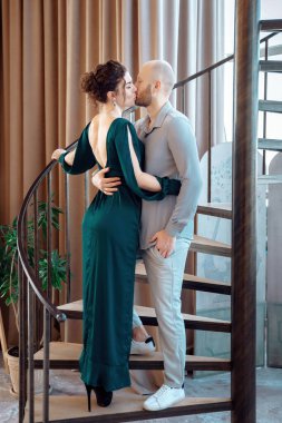 Lovely gently couple embracing with closed eyes standing on a spiral staircase. Passionate young man and woman are kissing. Attractive brunette woman in long evening dress and stylish bearded man