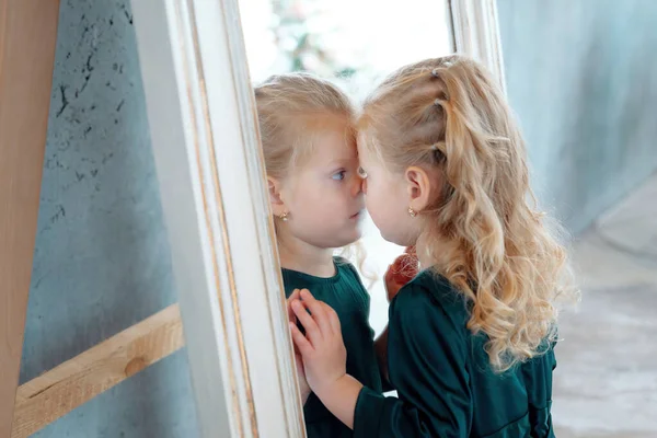 Una chica rubia linda se retuerce en el espejo. Divertido niño caucásico con vestido verde se mira en el espejo — Foto de Stock