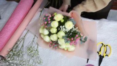 Çiçekçide buket oluşturma süreci, yakın çekim. Kadın elleri sarılı kağıt ve kurdeleyle çalışıyor. Çiçekçi bir buket gül yapar. Üst görünüm