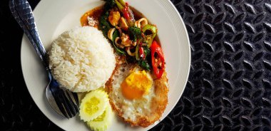 Fesleğen, kalamar, pirinç, salatalık ve kızarmış yumurta ile Tayland usulü beyaz tabakta fotokopi alanı ile karıştırılmış. Siyah paslanmaz çelikten arka planda ünlü ve baharatlı yiyecekler..