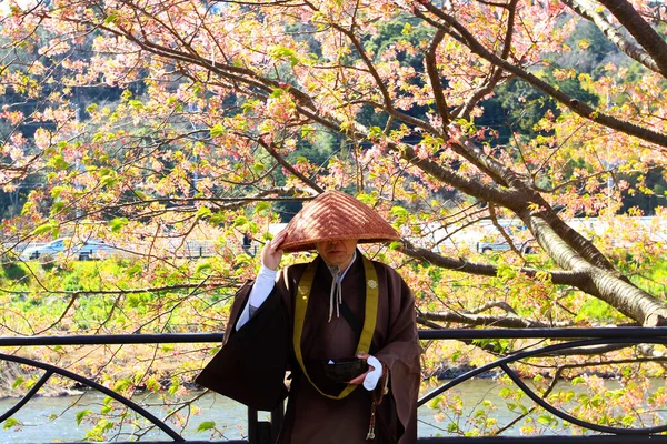 Japanese monk brown uniform, wearing straw hat and holding bowl for people donate money, food and general use with lake and sakura or pink flower tree background. Religion, belief and Beauty of natura