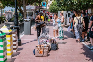 Sokakta bir sürü turisti olan bir sokak satıcısı kızlar için sahte çantalar satıyor. Popüler markalar da sahte çantalar. Marbella, Malaga / İspanya - 21 Temmuz 2022