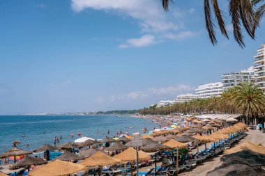 Şehir plajında deniz kıyısı boyunca bir sürü turist ve yaya caddesinin olduğu manzara. Plaj saman şemsiyeleri ve güverte sandalyeleri MARBELLA, MALAGA / SPAIN - 21 Temmuz 2022
