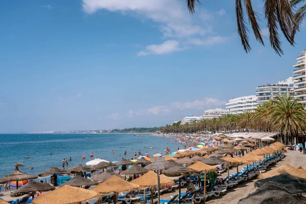 Şehir plajında deniz kıyısı boyunca bir sürü turist ve yaya caddesinin olduğu manzara. Plaj saman şemsiyeleri ve güverte sandalyeleri MARBELLA, MALAGA / SPAIN - 21 Temmuz 2022