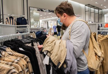 Sakallı ve tıbbi maskeli genç bir adam giyim mağazasından pantolon seçiyor.