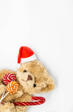 Bir kulağında kırmızı Noel Baba şapkası olan oyuncak oyuncak ayıyla dikey fotoğraf. Elinde baston şeker ve patilerinde iki çizgili lolipop var. Beyaz arkaplan, kopyalama alanı. Noel Kavramı.