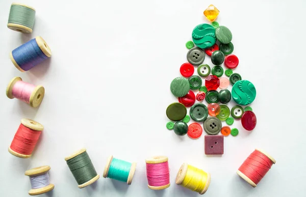 Düğmelerden yapılmış bir Noel ağacı, yeşil, kırmızı renk. Ahşap makaralı çok renkli iplikler. Beyaz arkaplan, kopyalama alanı. Yaratıcı Noel kavramı.