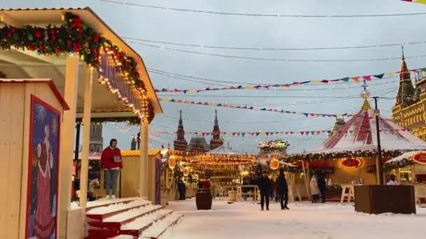 モスクワ ロシア 2021年12月 レッドスクエアのクリスマスフェア カルーセル メリーゴーランド 歩行者や照明スタンドでお祝いの市場販売 雪が降っている — ストック動画