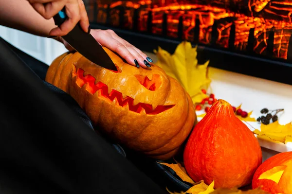 Una bruja en un impermeable con un cuchillo talla una calabaza para Halloween en casa, sobre el fondo de la chimenea — Foto de Stock