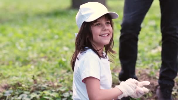 Side view video van een lachend meisje draagt witte pet en t-shirt fixes handschoenen terwijl het verzamelen van de vuilnis buiten. — Stockvideo