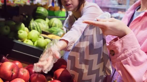 Ibu dengan putrinya mengenakan sarung tangan di toko memilih apel dan menempatkan mereka ke dalam tas belanja berdiri di dekat departemen buah. — Stok Video