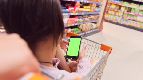 Zadní pohled video dívky sedí v nákupním vozíku se zkříženýma nohama drží smartphone v rukou se zelenou obrazovkou a matka tlačí vozík chůzi mezi regály v obchodě. — Stock video