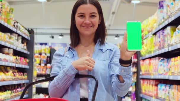 Close up video of a happy smiling young woman standing among the shelves in the store holding in her hands a grocery basket and the phone with green screen. — Stock Video