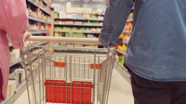 Powrót widok wideo z parą spacerujących wśród półek w sklepie trzymających wózek spożywczy razem, koncepcja zakupów. — Wideo stockowe