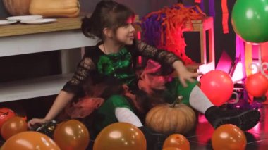 Cadılar Bayramı elbisesi giymiş güzel bir kız renkli balonların ve balkabaklarının arasında oturuyor ve onları yükselten balonlarla oynuyor.. 