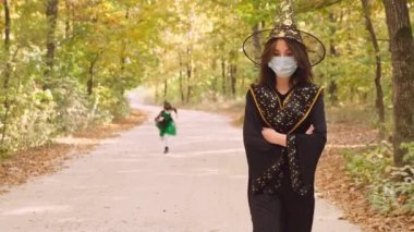 Cadılar Bayramı partisi için siyah elbiseli genç bir bayanın videosunu kapat. Ormanda tıbbi maske takmış küçük bir kız koşuyor..