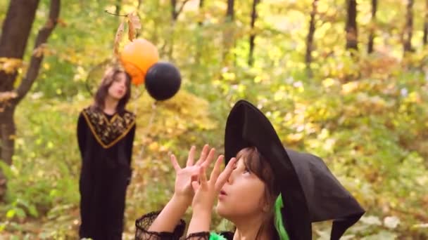 Две девушки в осеннем лесу, одетые в хэллоуинскую одежду, одна колдует с листьями деревьев, а другая смотрит, держа в руках два шара. — стоковое видео