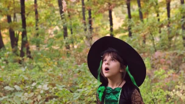 Magician girl wearing Halloween dress and hat blowing at the leaves to raise them up making witchcraft outdoors. — Stock Video