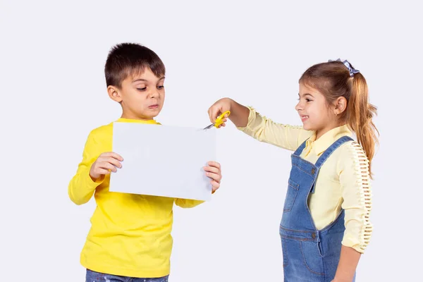 Foto av två barn, pojke håller vitt papper i sina händer och flicka försöker skära papper med sax gul färg, pojken ser med oro. — Stockfoto
