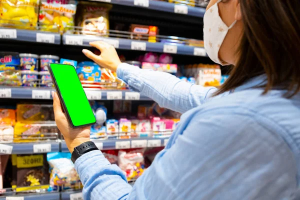 Καλλιεργημένη φωτογραφία νεαρής γυναίκας με μάσκα προσώπου χρησιμοποιώντας κινητό τηλέφωνο και αγοράζοντας είδη παντοπωλείου στο σούπερ μάρκετ κατά τη διάρκεια πανδημίας ιών. — Φωτογραφία Αρχείου