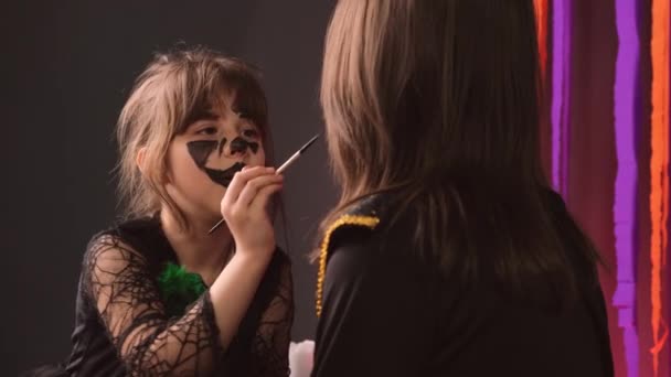 Dívka s černým akvagrim vzor na její tváři malování na její sestře tvář pomocí štětce připravuje na Halloween party dnes. — Stock video