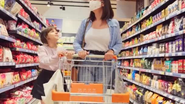 Küçük kızıyla birlikte tıbbi maske takan bir kadın marketteki rafların arasında yürürken market arabasıyla alışverişe çıkıyor ve kızı eline dokunurken bir şey söylemek istiyor.. — Stok video