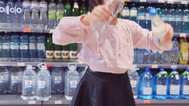 Close up video veselá dívka drží v ruce dvě láhve vody a tanec v obchodě přes láhve na poličkách pozadí. — Stock video