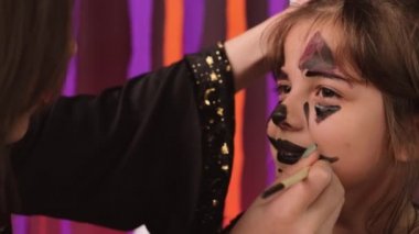 Küçük kıza çizilen fırça ve siyah renkli boyaları kullanan genç bir kadın, Cadılar Bayramı için maske takıyor. Yüksek kalite 4k görüntü