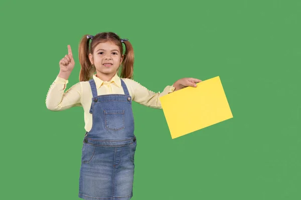 Η μαθητριούλα δείχνει τους αντίχειρες στο πράσινο φόντο κρατώντας το κίτρινο χαρτί στο χέρι της και δείχνοντας με το δάχτυλό της ψηλά, δίνοντας προσοχή.. — Φωτογραφία Αρχείου