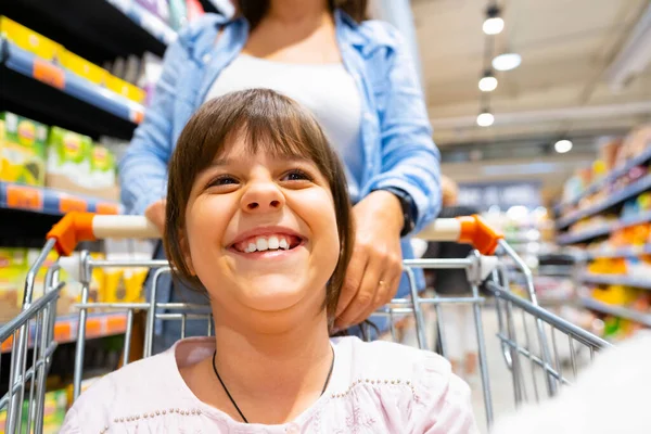 Κορίτσι κάθεται στο καλάθι αγορών χαμογελώντας ευρέως ψώνια στο κατάστημα που διέρχεται μεταξύ των ραφιών με πολλά προϊόντα. — Φωτογραφία Αρχείου