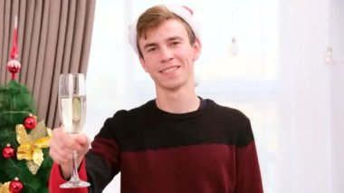 Noel Baba şapkalı genç adam elinde şampanya bardağıyla yılbaşı ağacının yanında duruyor ve kameraya doğru koşuyor..